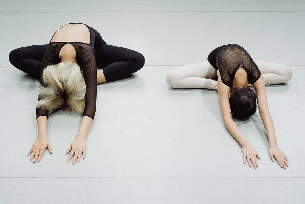 Two female dancers performing a butterfly stretch in a dance studio