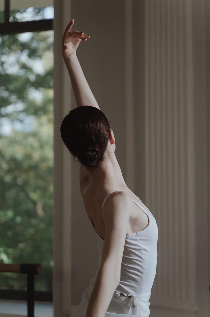 A female dancer in a white leotard with arm extending upwards to highlight ballet technique